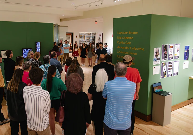 A diverse group of people gathered in an exhibition space, attentively listening to speakers in front of a green display wall featuring various artworks.