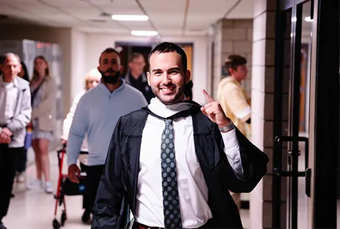 A smiling graduate in a cap and gown walks through a hallway, celebrating their accomplishment.