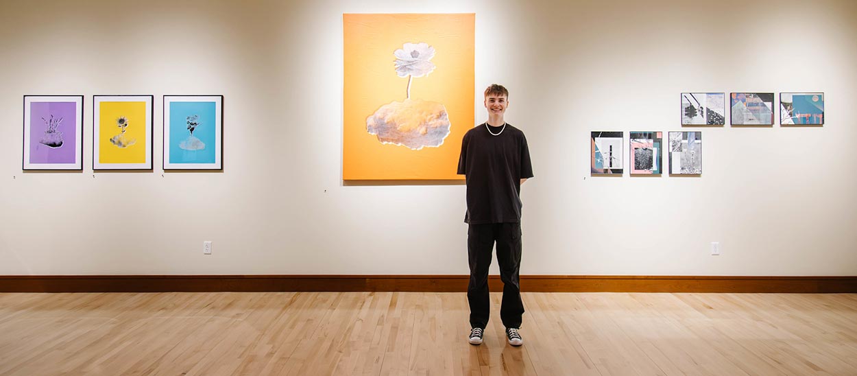 Student smiling standing in a gallery with his protfolio displayed in frames on the wall behind him