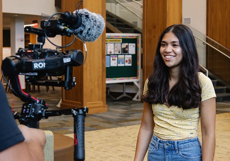 A young woman smiles while being interviewed on camera in a well-lit indoor setting.