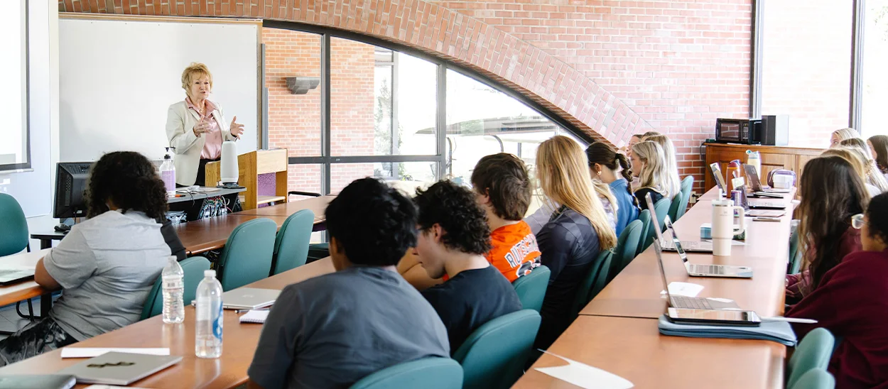 A woman delivers a presentation to a group of seated students in a modern classroom setting.