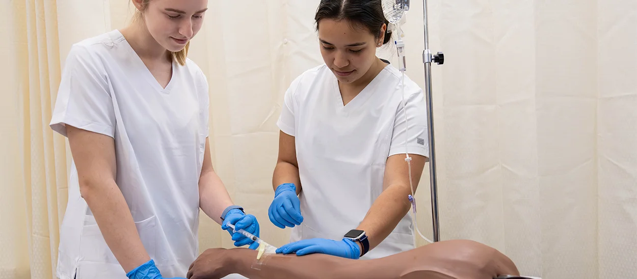 Two nursing students in scrubs practicing an intravenous catheter insertion on a medical training mannequin.