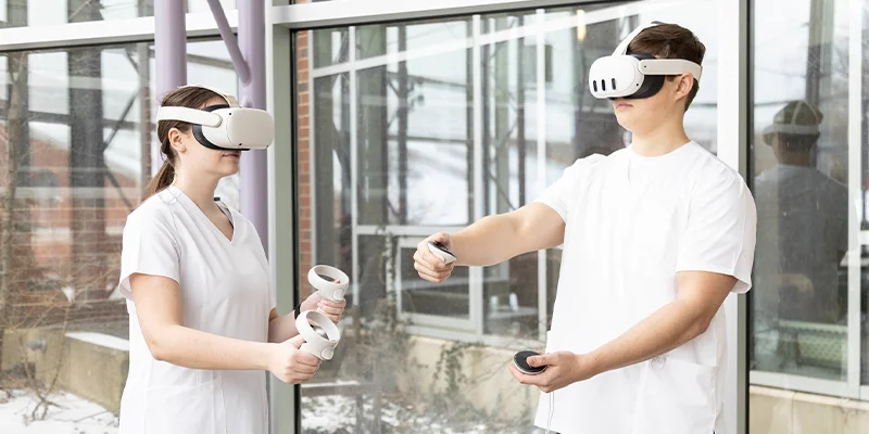 Two individuals in white scrubs are engaged in a virtual reality exercise, wearing VR headsets and holding controllers in a modern, bright space.