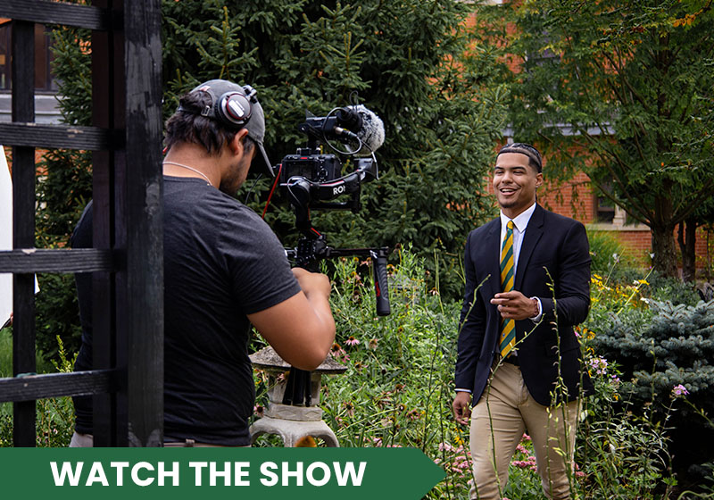 A man in a suit is being filmed outdoors, smiling at the camera while a crew member operates the filming equipment.