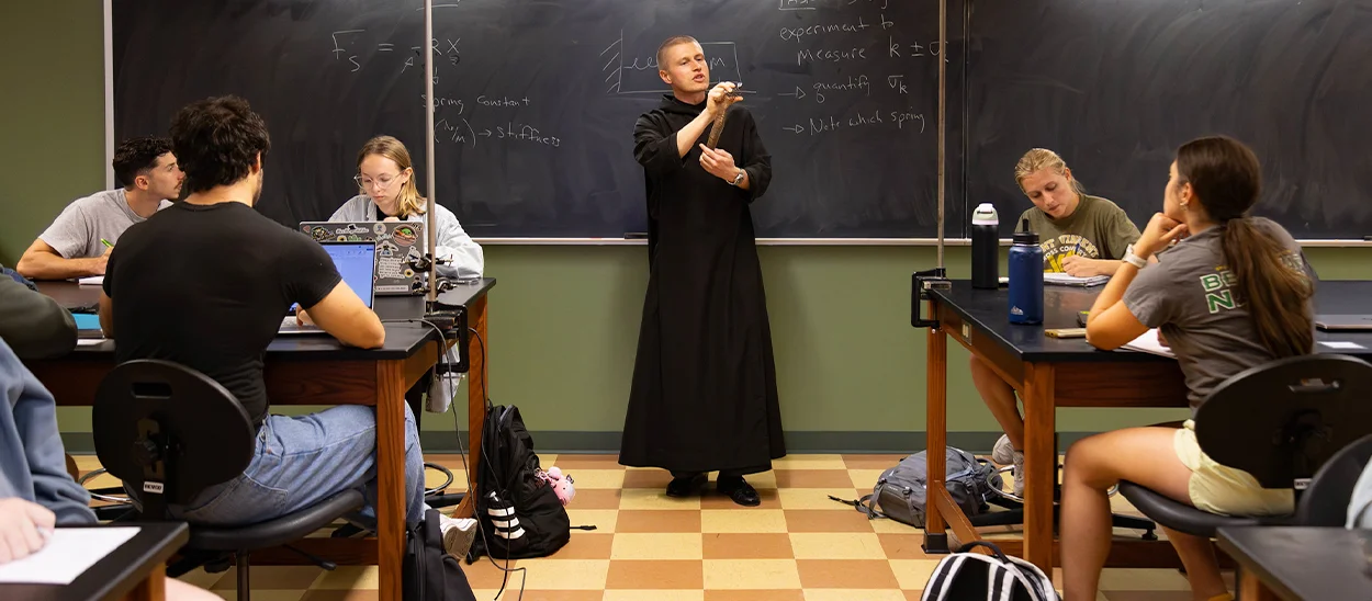 A professor in a black monk's robe teaching a classroom of students, with mathematical equations written on the chalkboard.