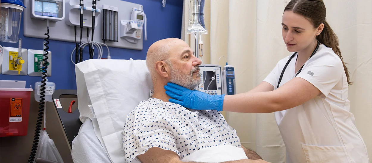 A healthcare student in scrubs examines a patient in a hospital bed.