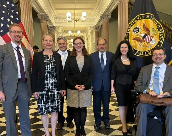 Group photo of professionals with Aileen Mooney in the middle standing in a formal setting, featuring an American flag and a presidential seal in the background.