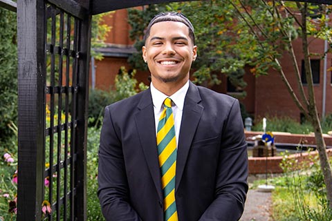 A young man in a black suit and yellow-green striped tie stands smiling in a garden setting.