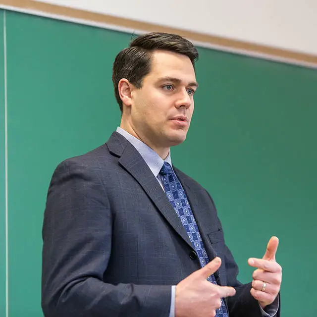 Dr. Jerome Foss gestures while speaking in front of a green chalkboard.