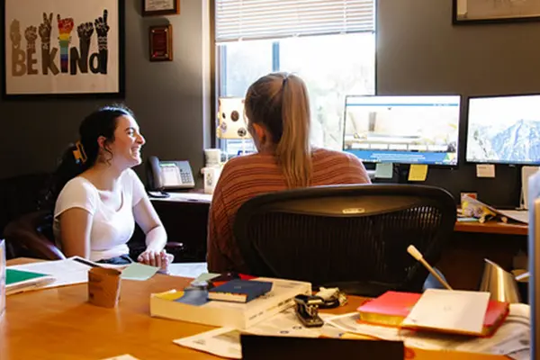 Two women engaged in discussion at a desk with computers and paperwork, highlighting a collaborative workspace.