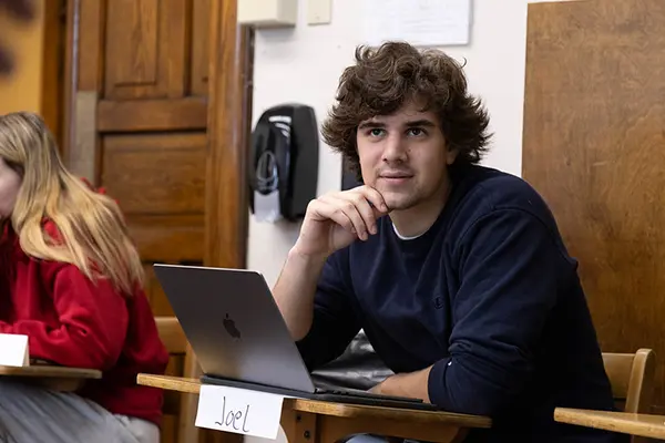 A student named Joel sits at a desk in a classroom, looking thoughtful while using a laptop.