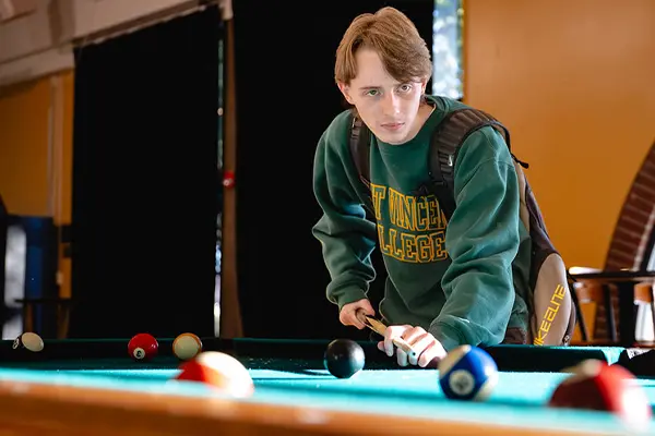 A student wearing a college sweatshirt concentrates while preparing to take a shot at a pool table.