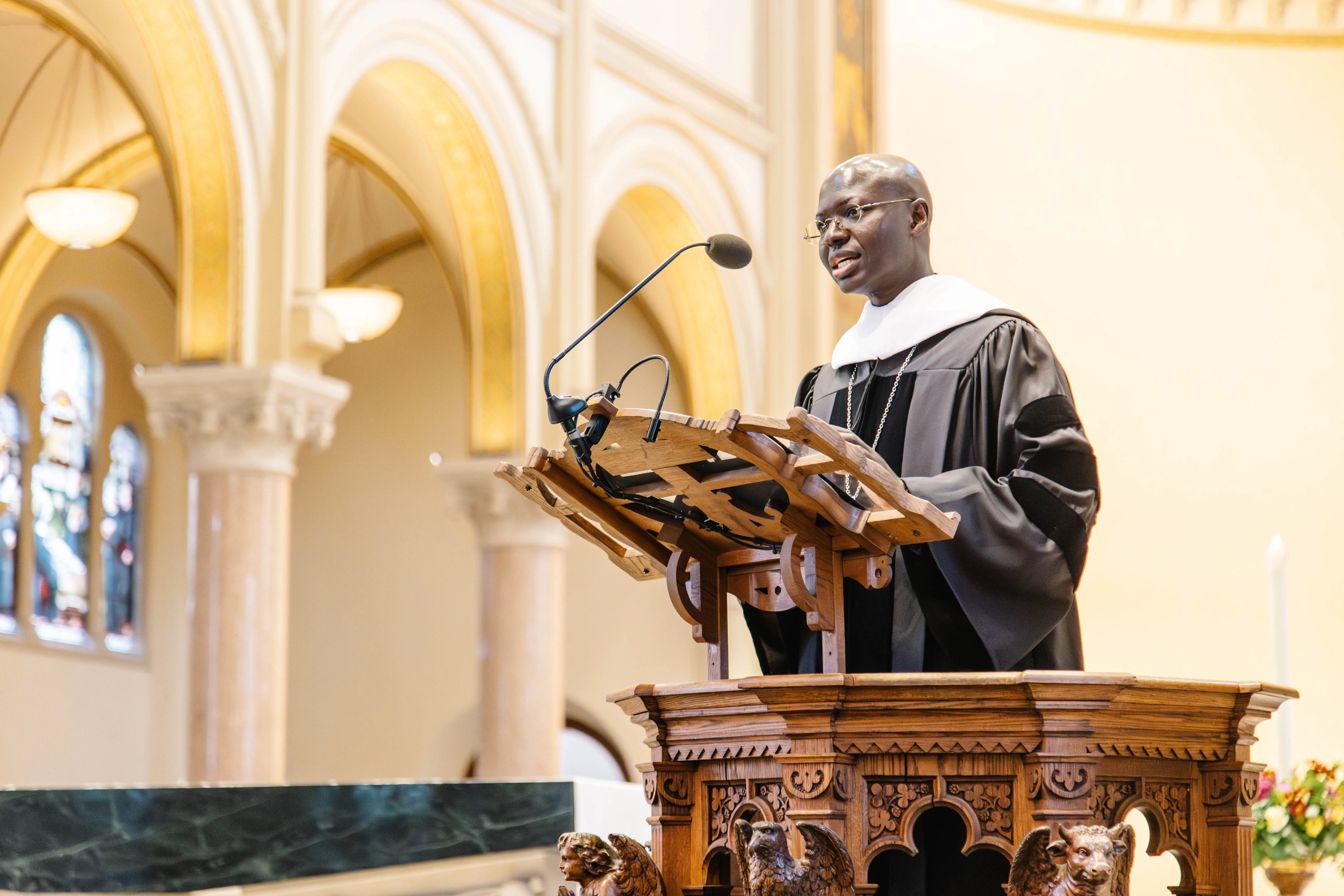 A man in academic robes delivers a speech from a beautifully carved wooden lectern inside a church.