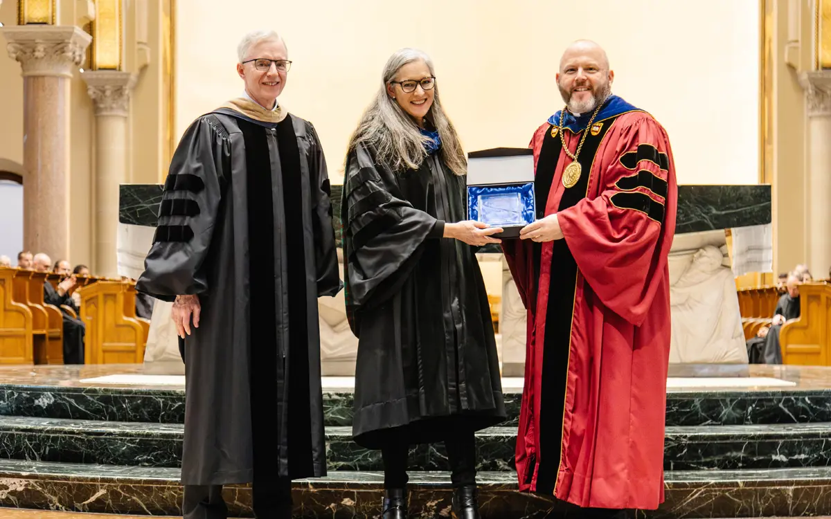 A woman in academic regalia receives an award from two male figures in formal attire during a ceremony.