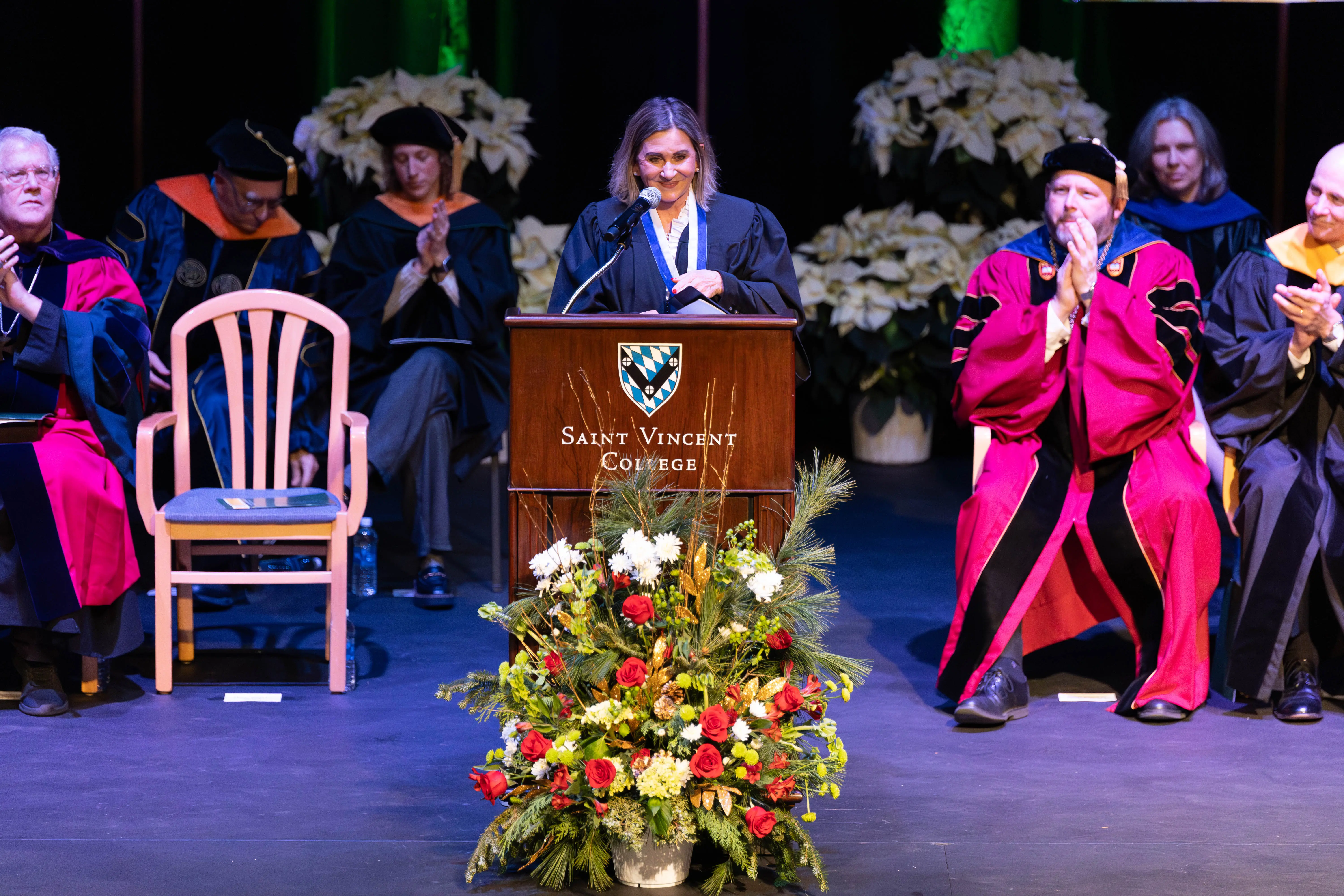 A graduation ceremony scene featuring a female speaker at a podium with the Saint Vincent College logo, surrounded by faculty in academic regalia and a floral arrangement in front.