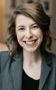 A smiling woman in a gray blazer, with shoulder-length wavy hair, exuding a friendly and approachable demeanor.