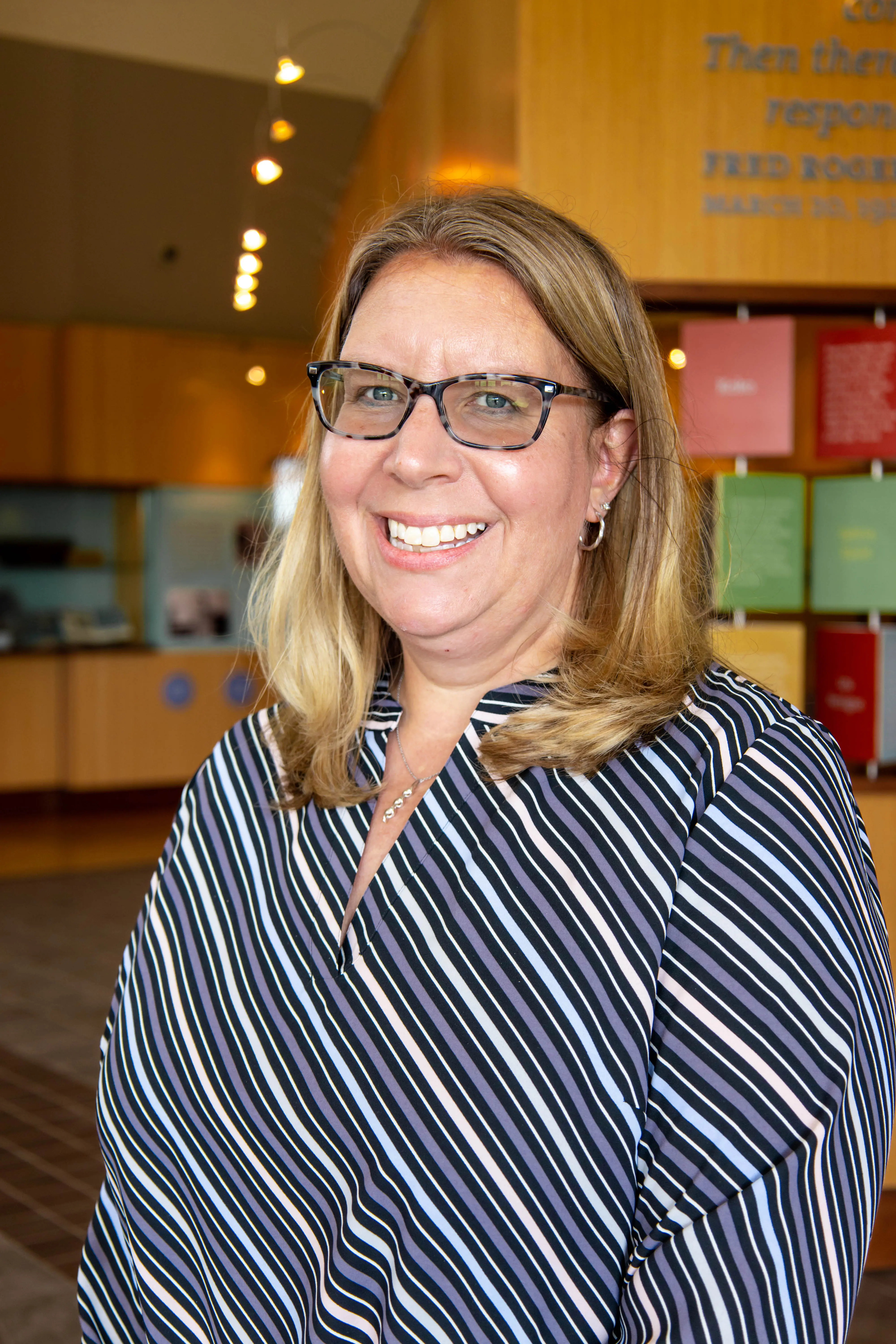 A smiling woman with glasses wearing a striped blouse stands in a welcoming indoor space.