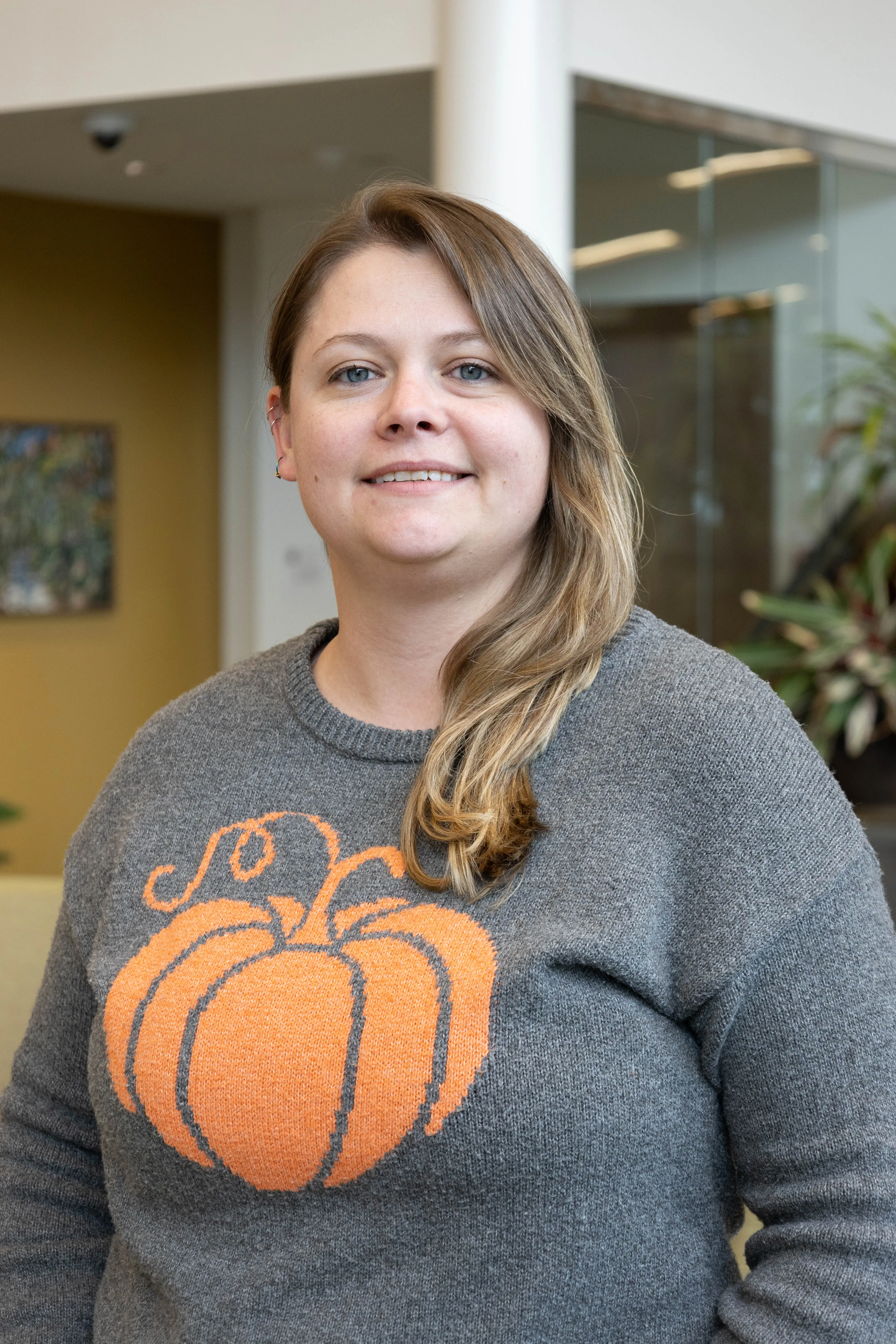 Woman wearing a grey sweater with an orange pumpkin design, smiling while standing in a modern indoor setting.