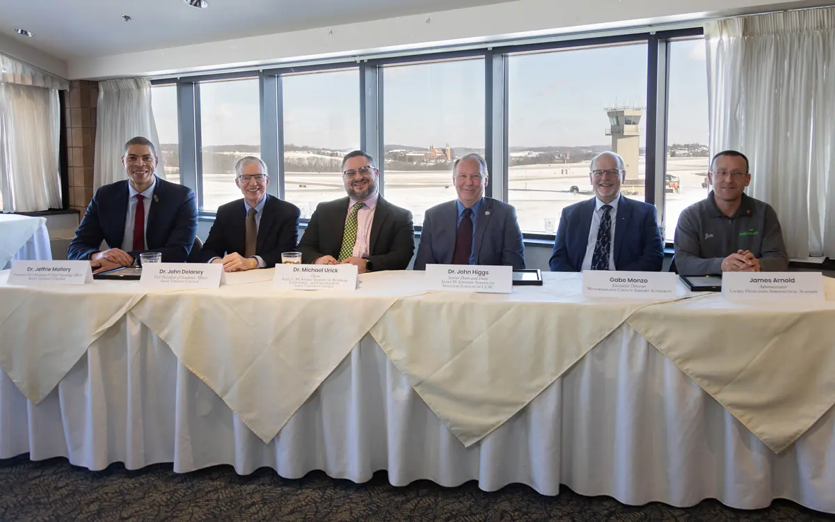 Group of six professionals seated at a table during a meeting with a view of an airport runway in the background.