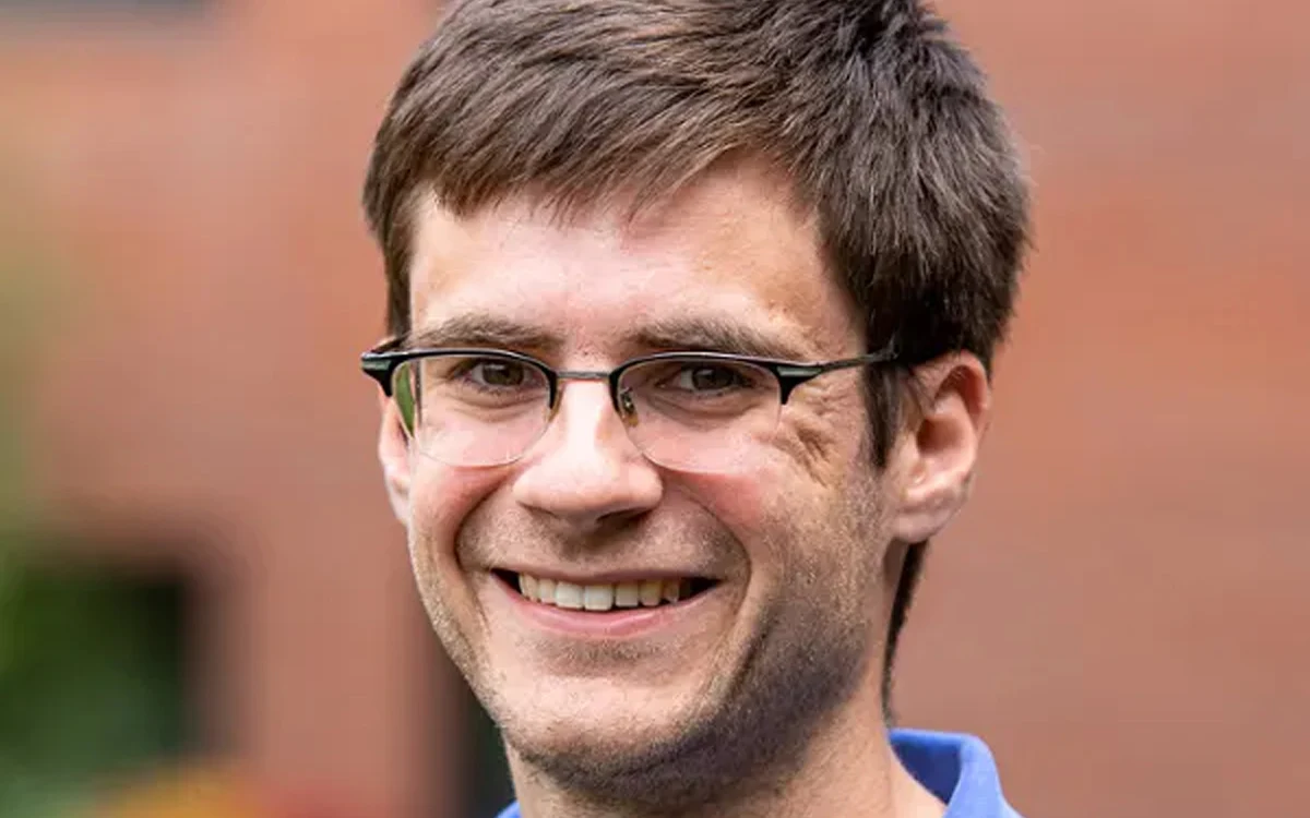 A smiling man with glasses and short brown hair, wearing a blue shirt.