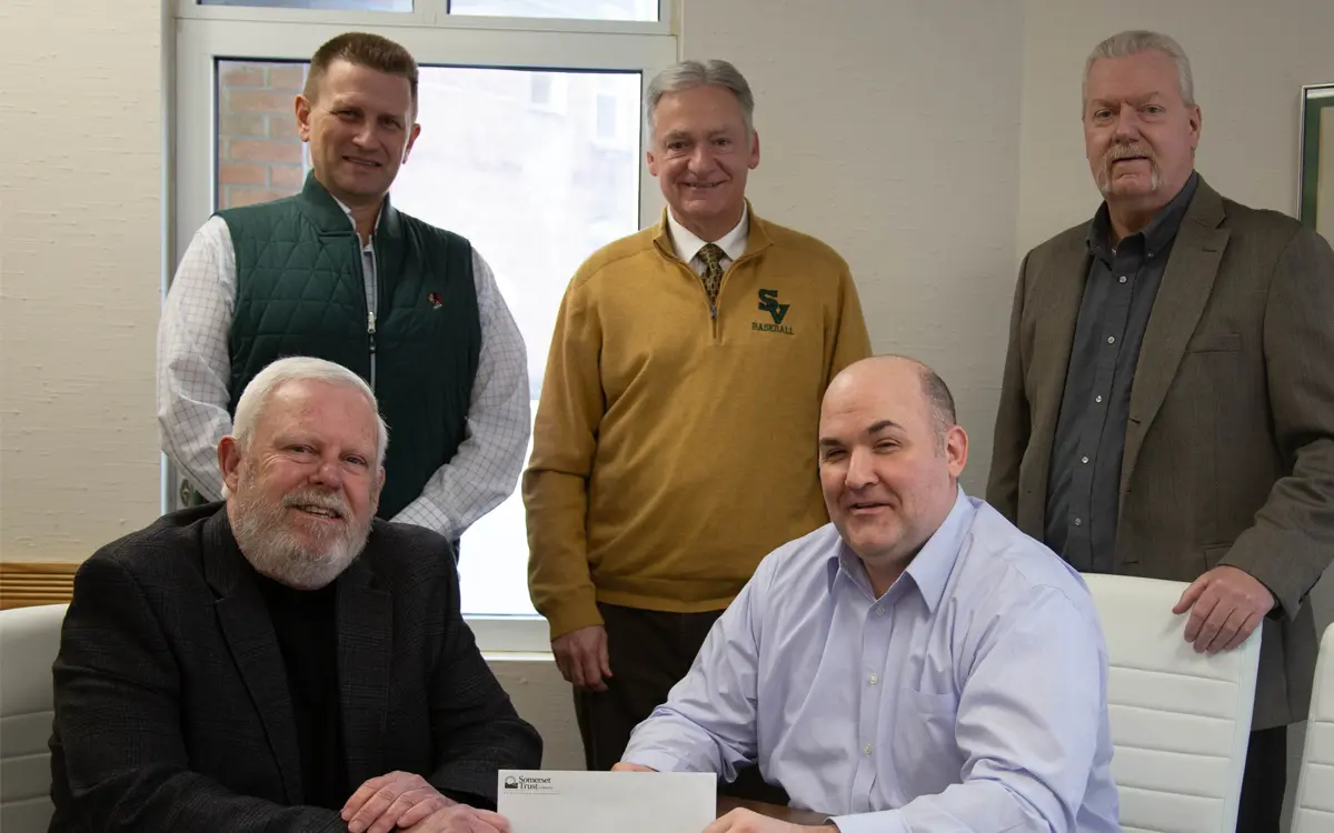 Group of five men, including one seated at a table signing documents, with two standing in the background and the others flanking them, discussing a partnership or agreement.