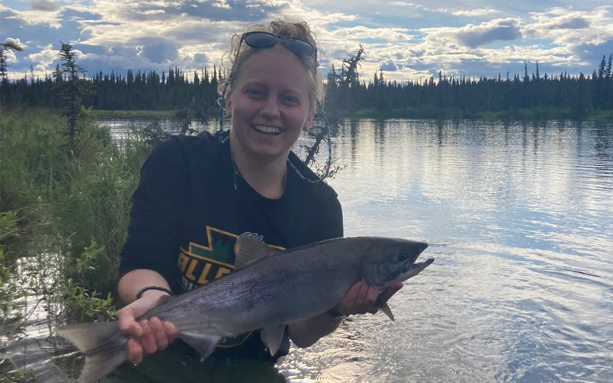 A smiling woman holds a large fish by a river, surrounded by lush greenery and a cloudy sky.