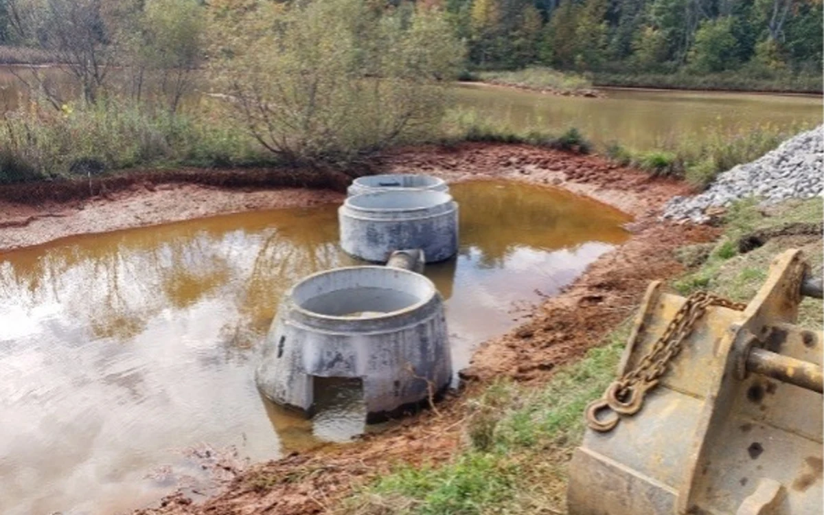 Two large concrete drainage structures partially submerged in a shallow, muddy pond surrounded by trees and vegetation.