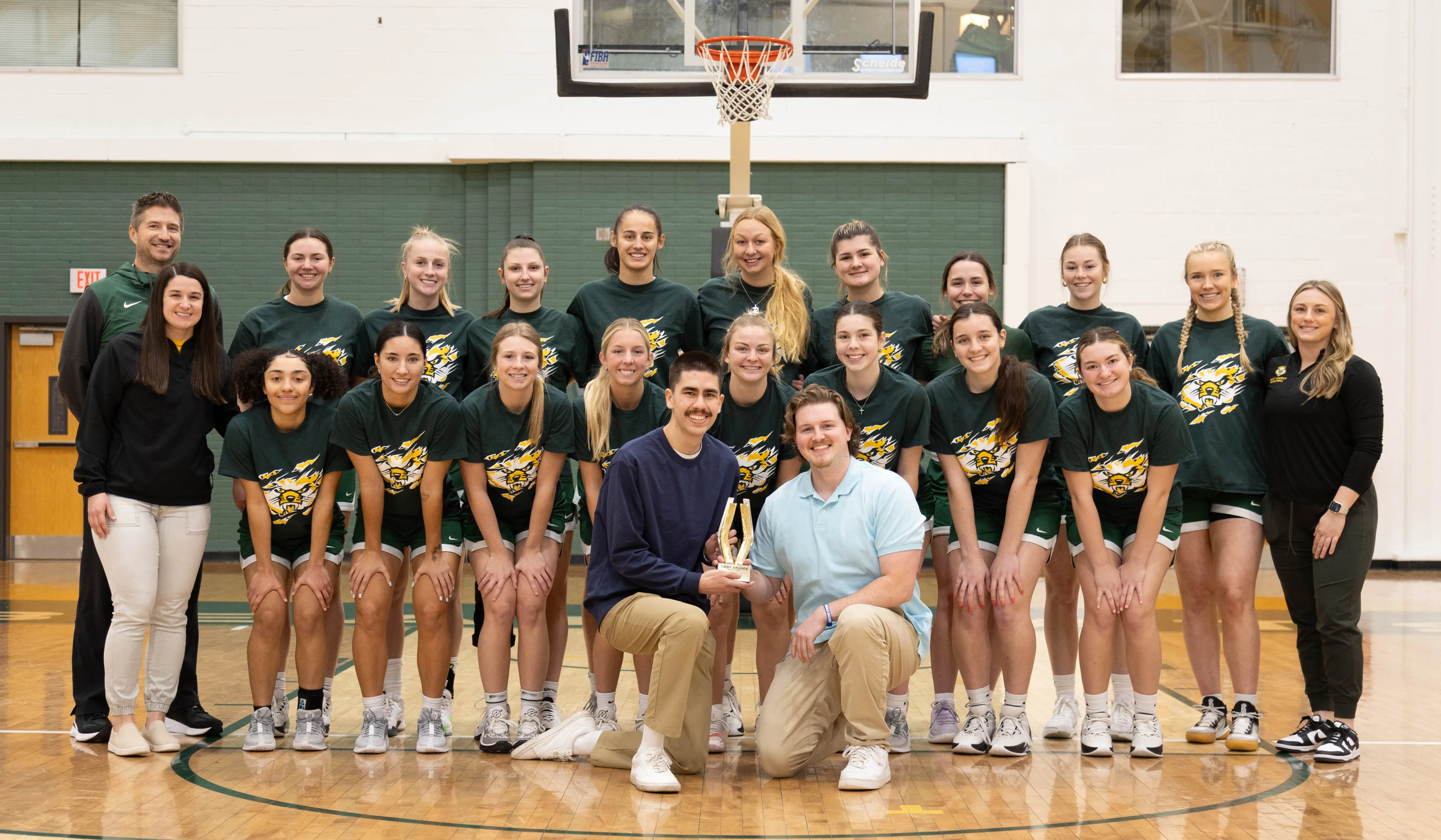 A group photo of a women's basketball team celebrating a trophy win, featuring players, coaches, and support staff in their uniforms.