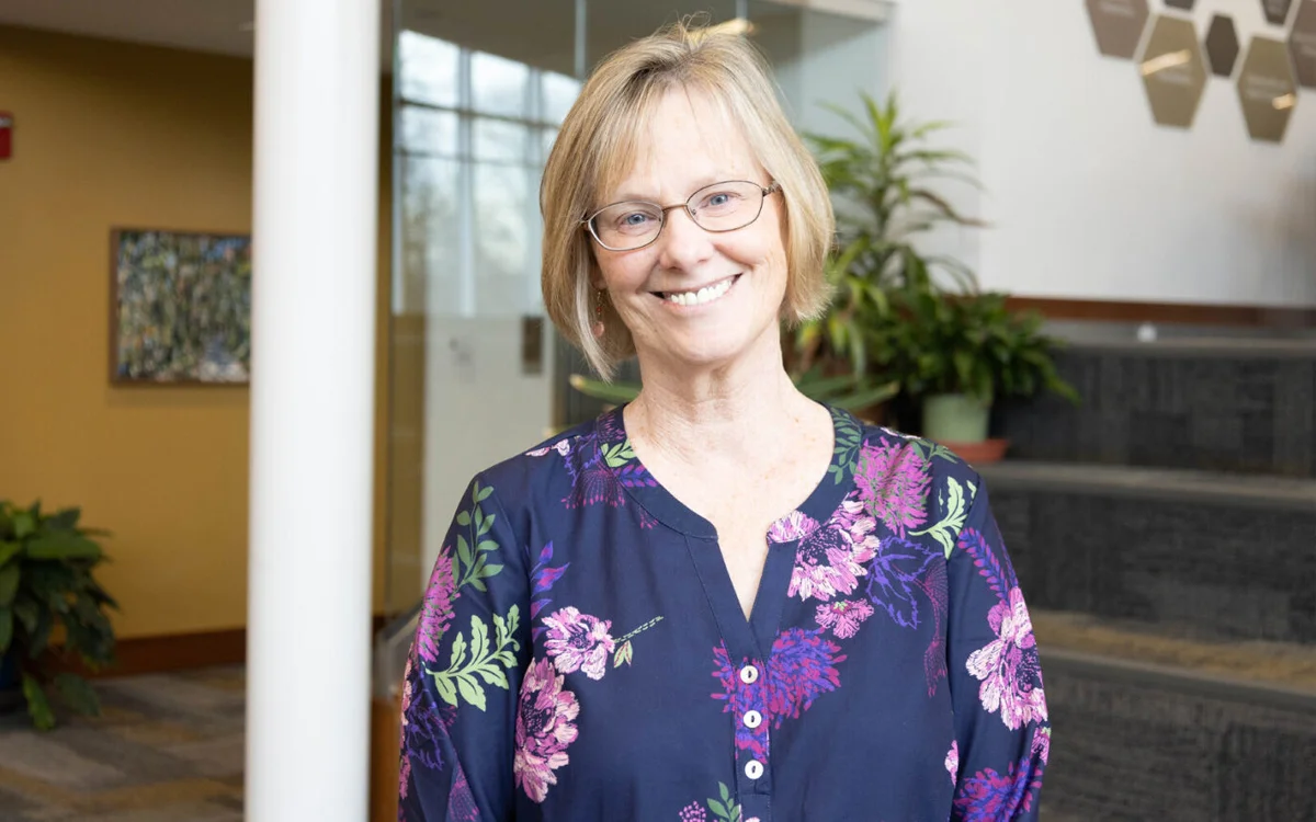 A smiling woman with short blonde hair and glasses, wearing a floral-patterned shirt, standing in a bright indoor space with plants in the background.