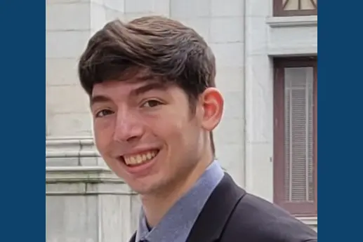 A young man with short brown hair smiles confidently while wearing a suit and tie, standing in front of a historic building.