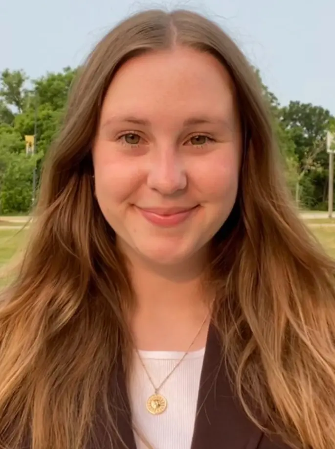 A smiling young woman with long hair wearing a blazer and a necklace, standing outdoors with greenery in the background.