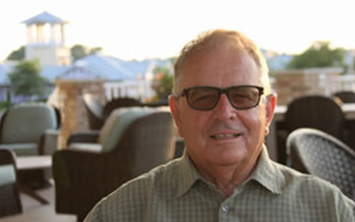 A smiling older man wearing glasses and a collared shirt, seated outdoors with a blurred background of chairs and buildings.