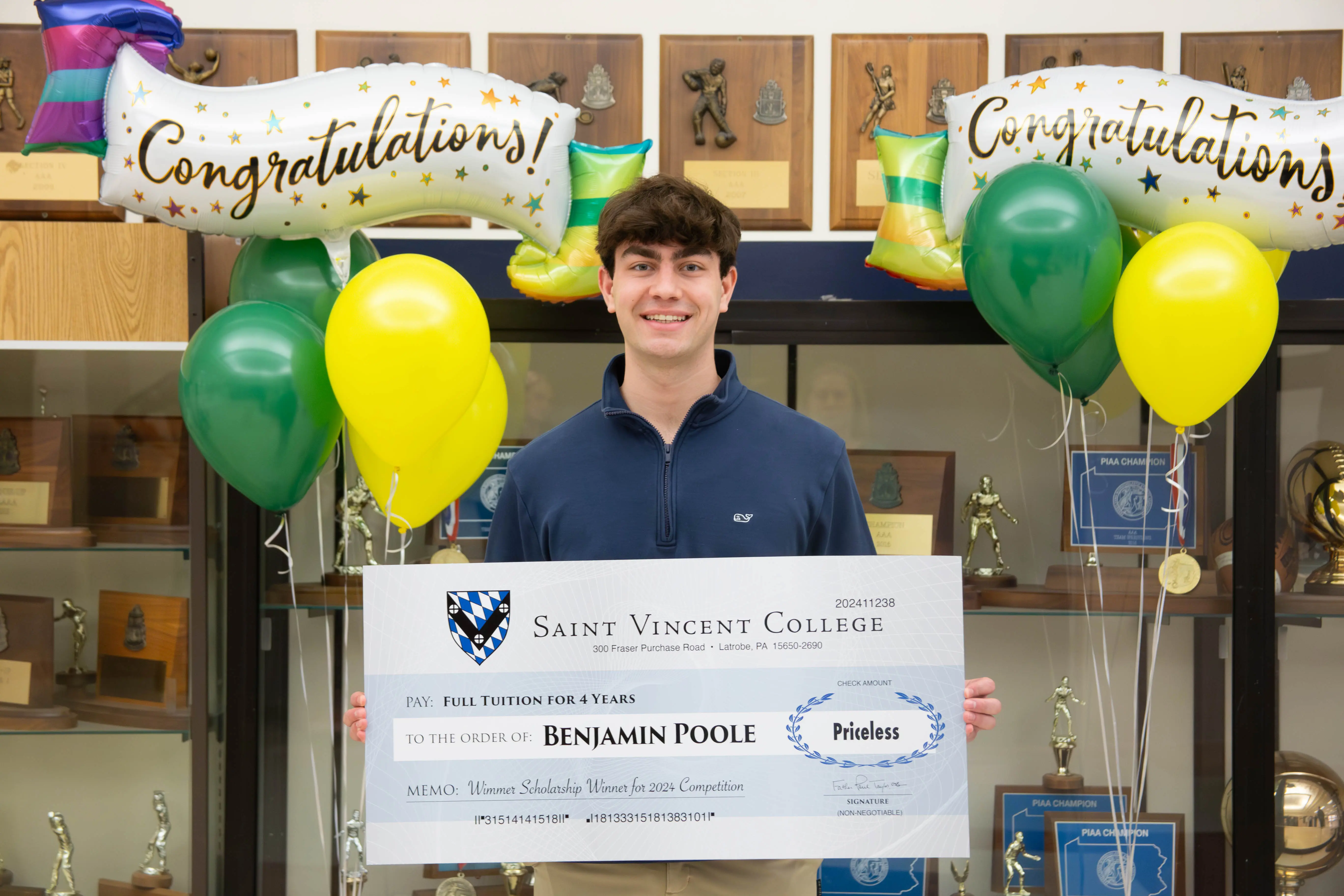 A student holding a large scholarship check from Saint Vincent College, surrounded by celebratory balloons and banners that read "Congratulations."