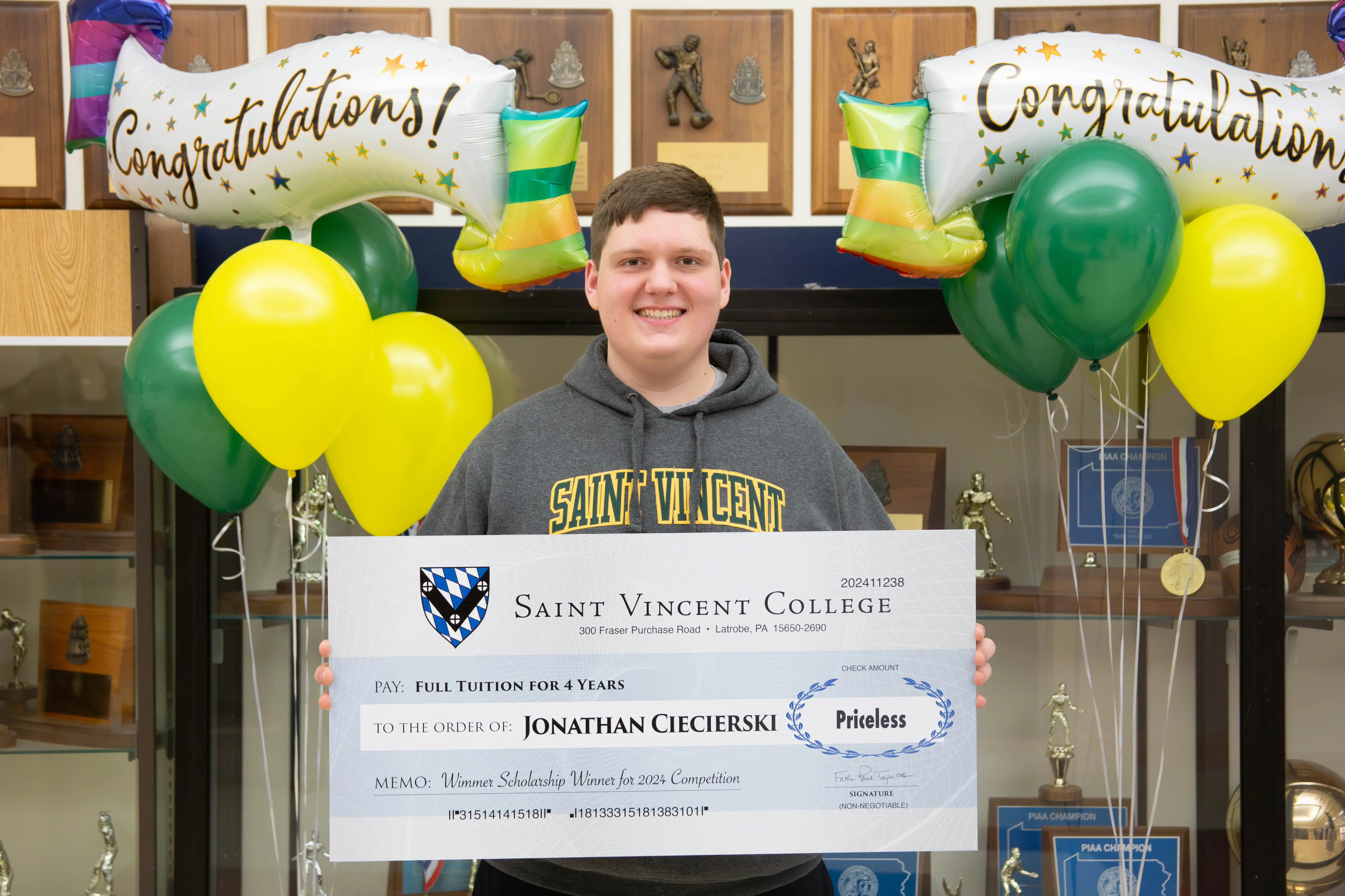 A student holding a large scholarship check for full tuition at Saint Vincent College, surrounded by balloons and congratulatory banners.