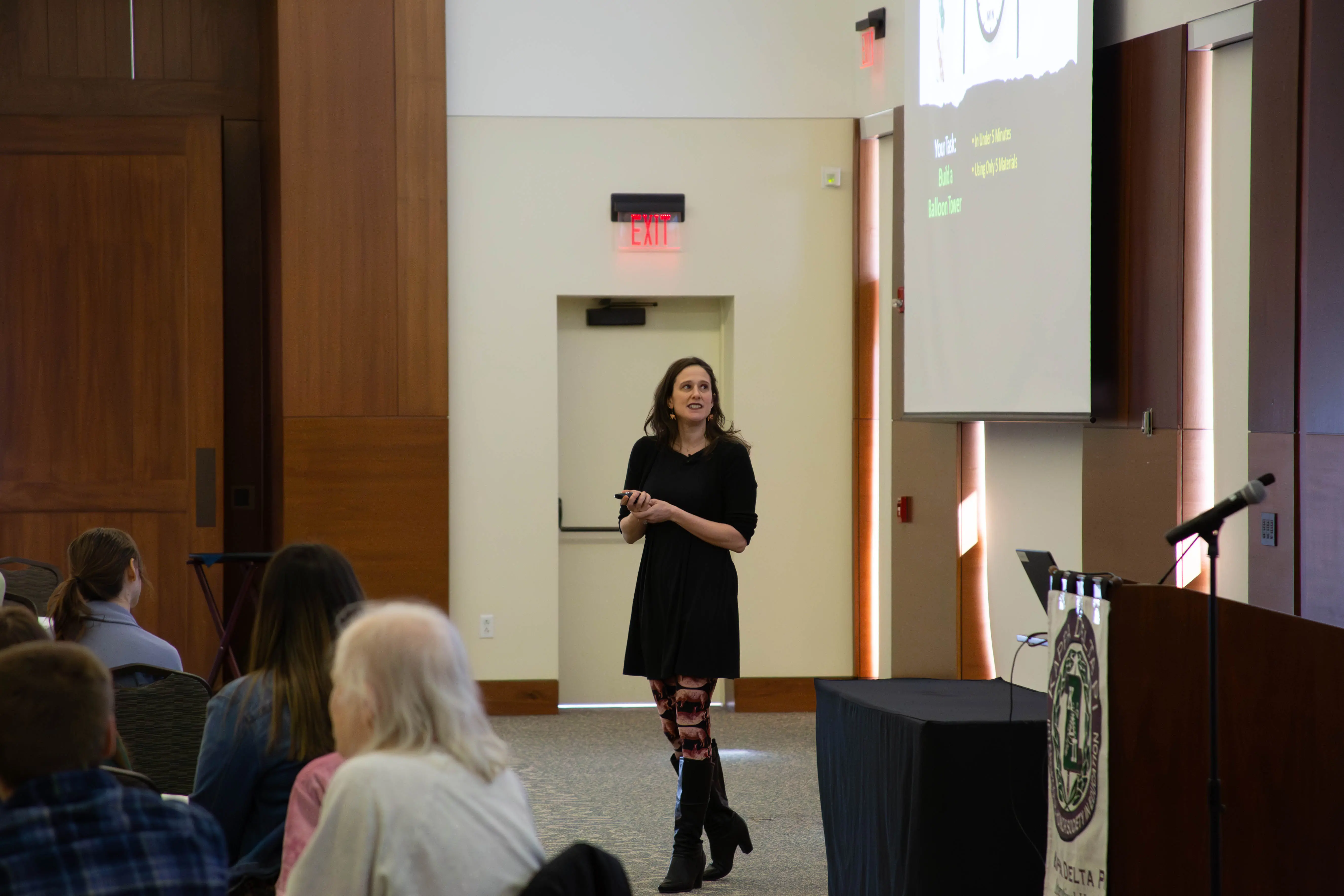 A speaker presenting to an audience in a conference room, with a projector screen displaying slides behind her.