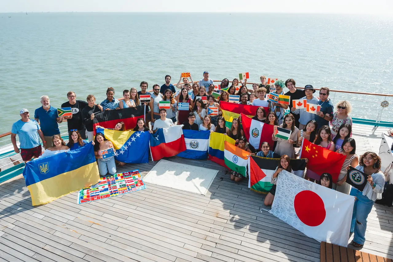 A diverse group of people holding various national flags, standing together on a deck by the water.