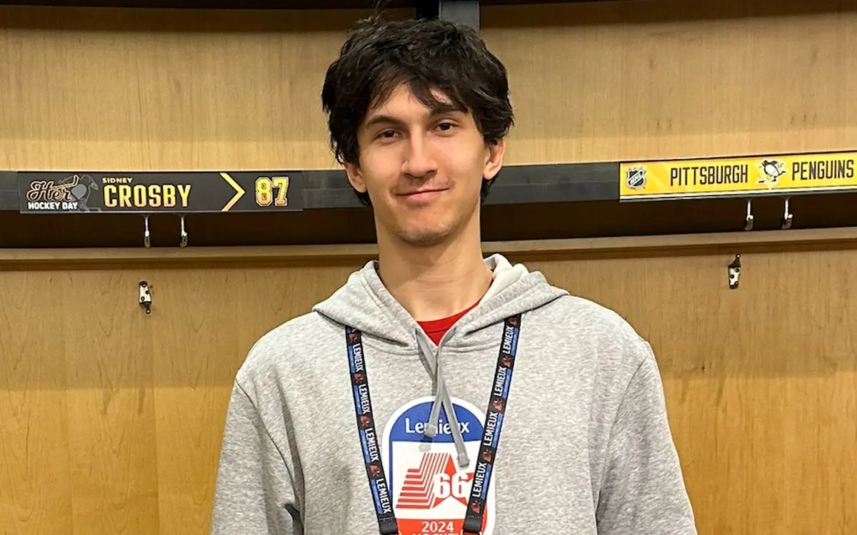 A young man wearing a gray hoodie stands in front of a hockey locker, showcasing his lanyard and a logo on his shirt.