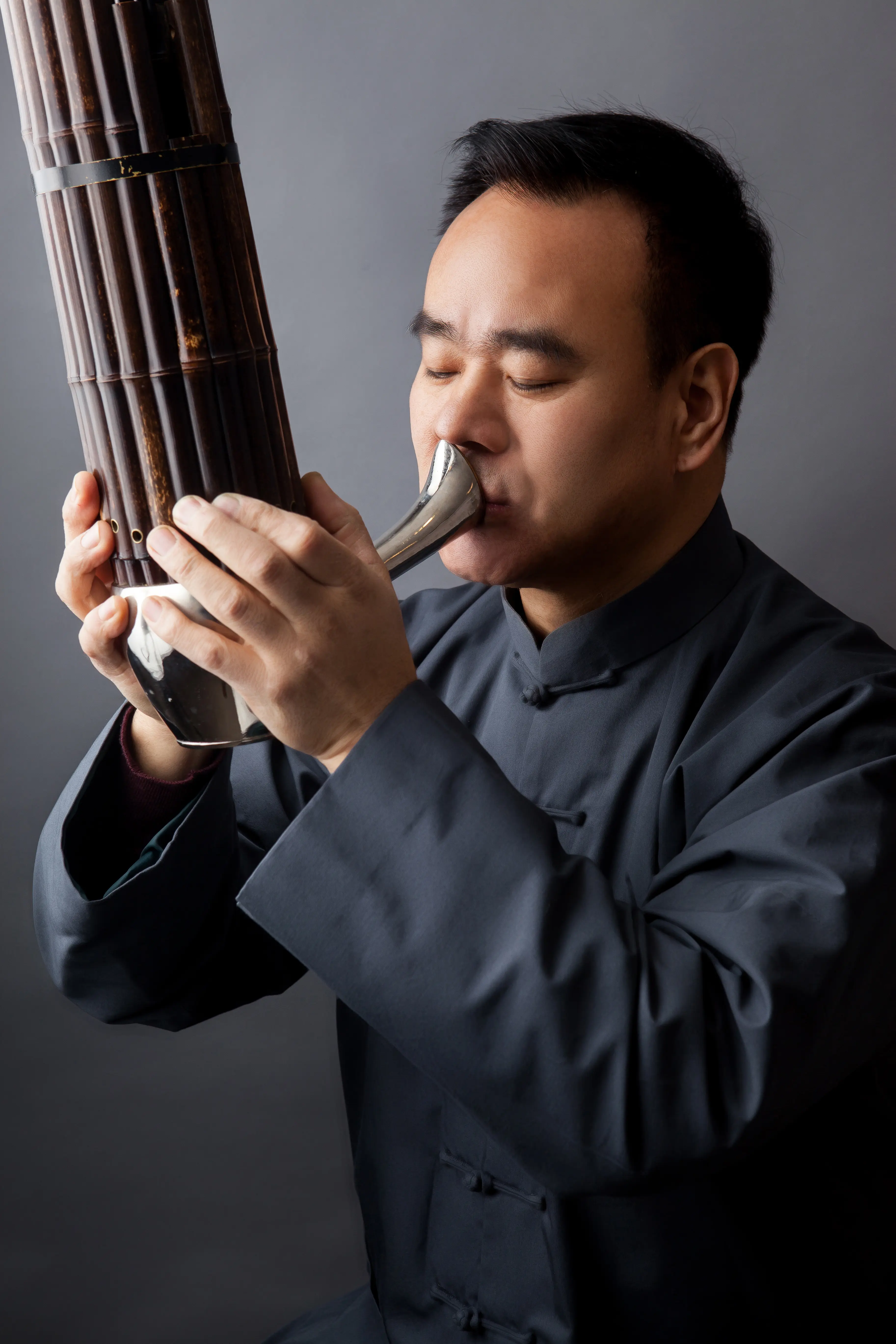 A musician playing a traditional wind instrument, the sheng, while wearing a dark traditional outfit.