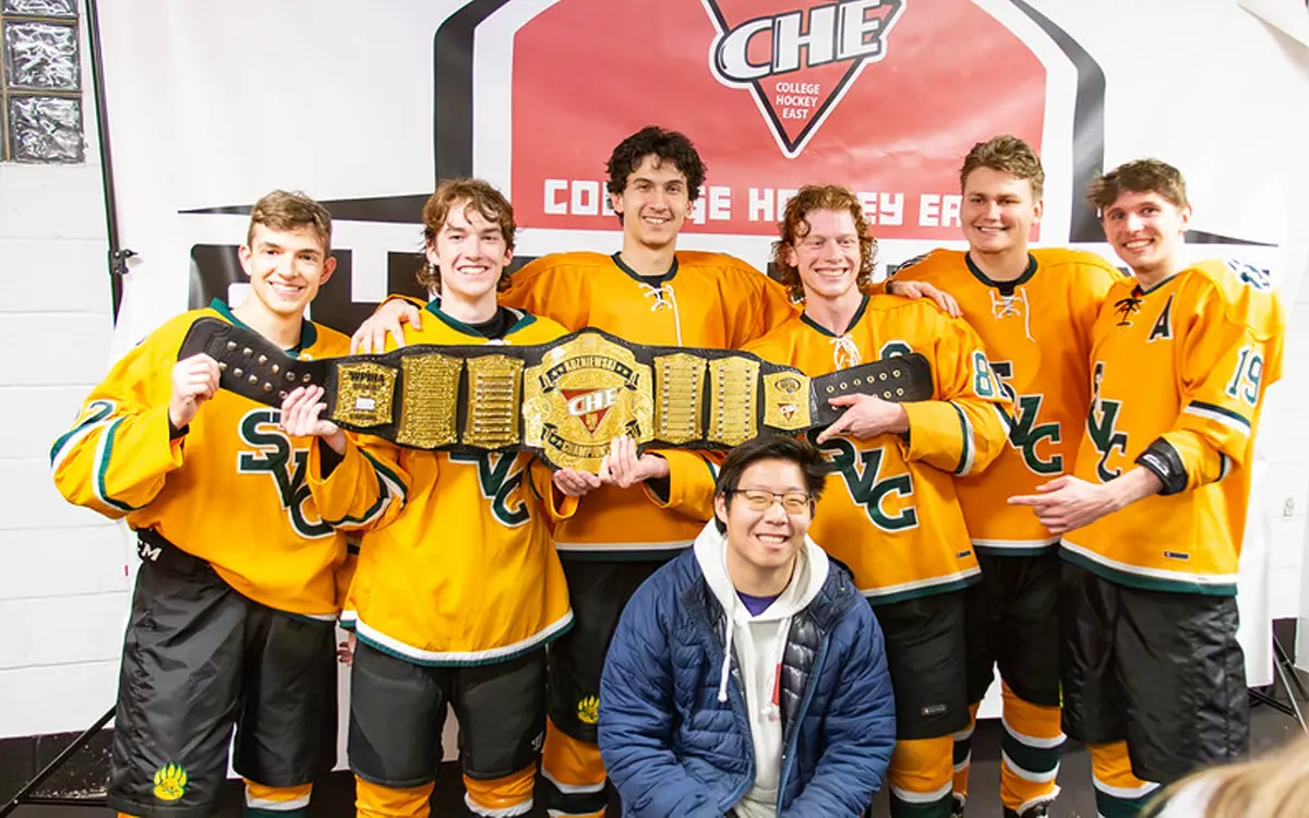 Group of college hockey players in orange jerseys holding a championship belt, celebrating their victory with a smiling fan.