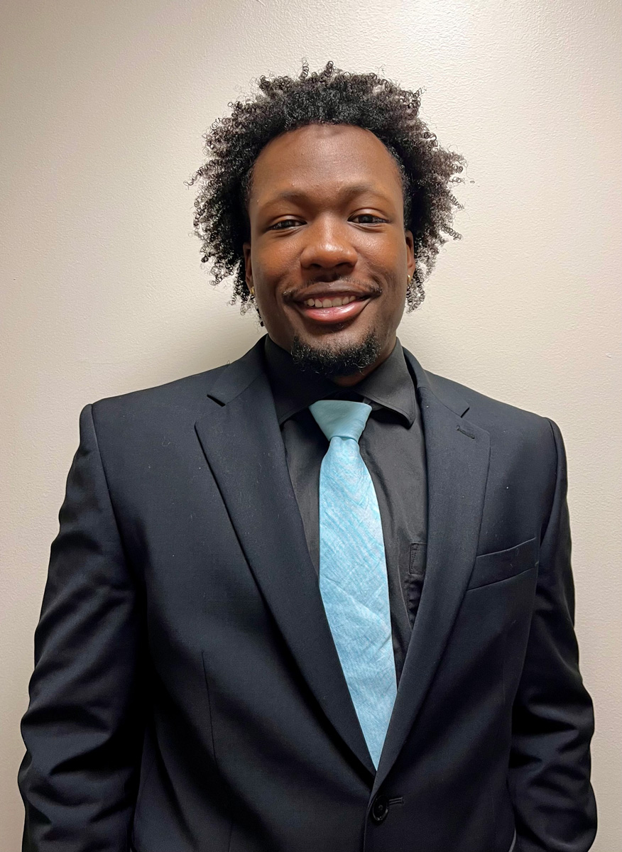 A man in a black suit and light blue tie, smiling against a neutral background.