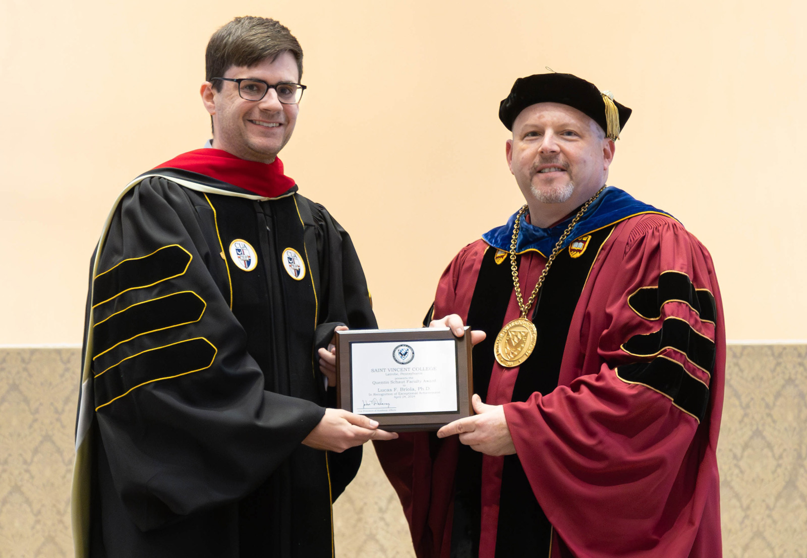 Lucas Briola and Father Paul in academic regalia are proudly holding the award smiling for the photo.