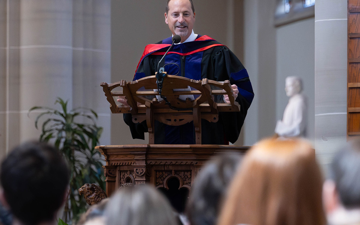 Daniel Lynch in academic regalia delivers a speech at a podium during a formal event.