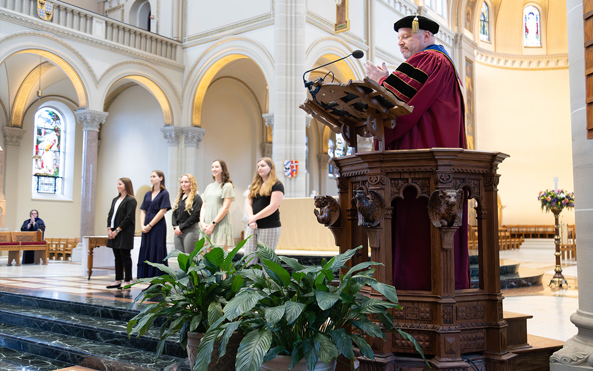 Father Paull in academic regalia delivers a speech at a ceremony, with students standing on stage in the background.