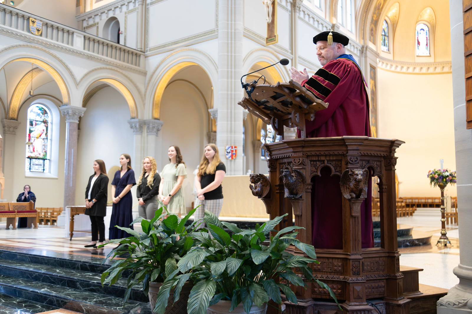 Father Paull in academic regalia delivers a speech at a ceremony, with students standing on stage in the background.