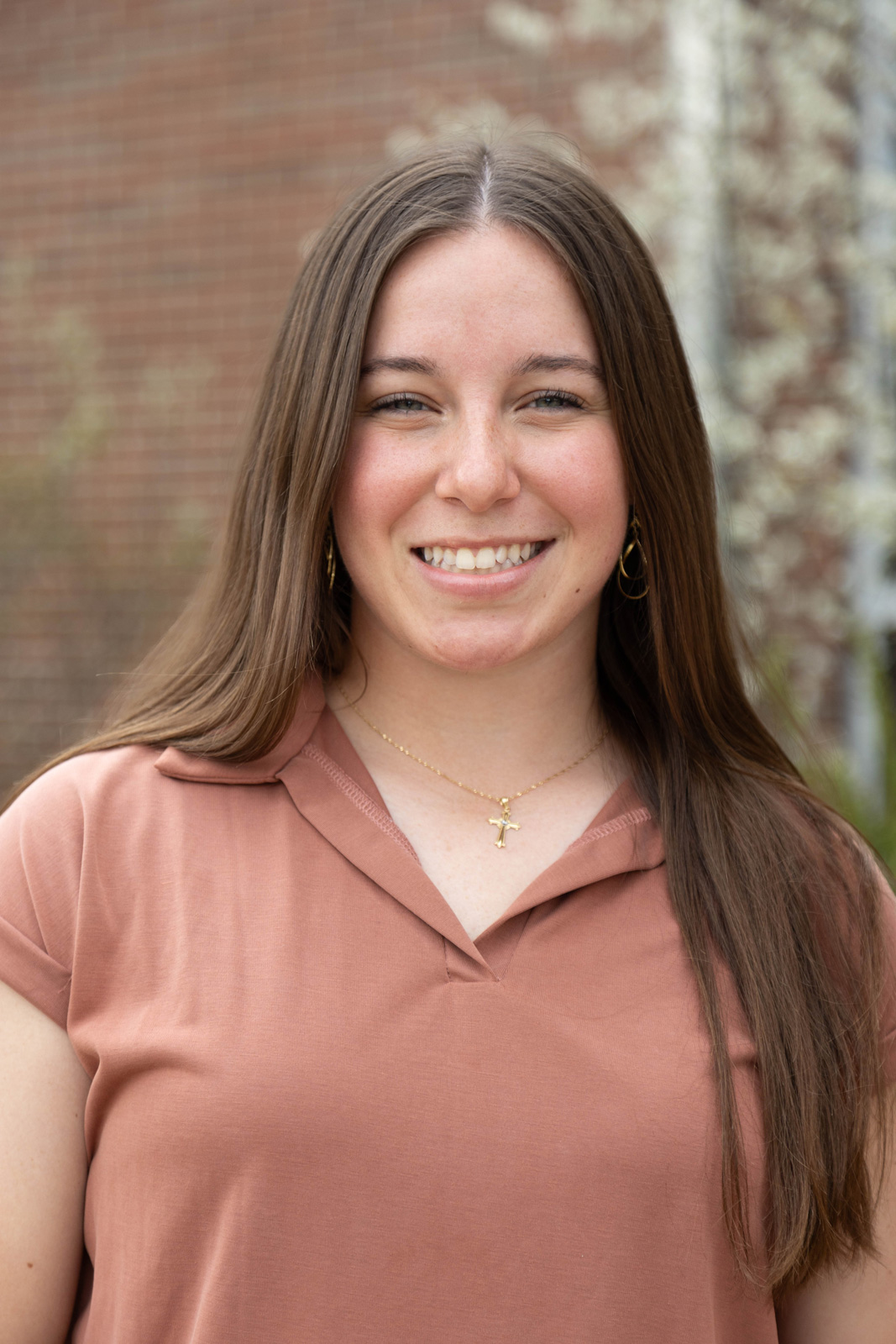 A smiling young woman with long hair, wearing a brown shirt and gold jewelry, stands outdoors.