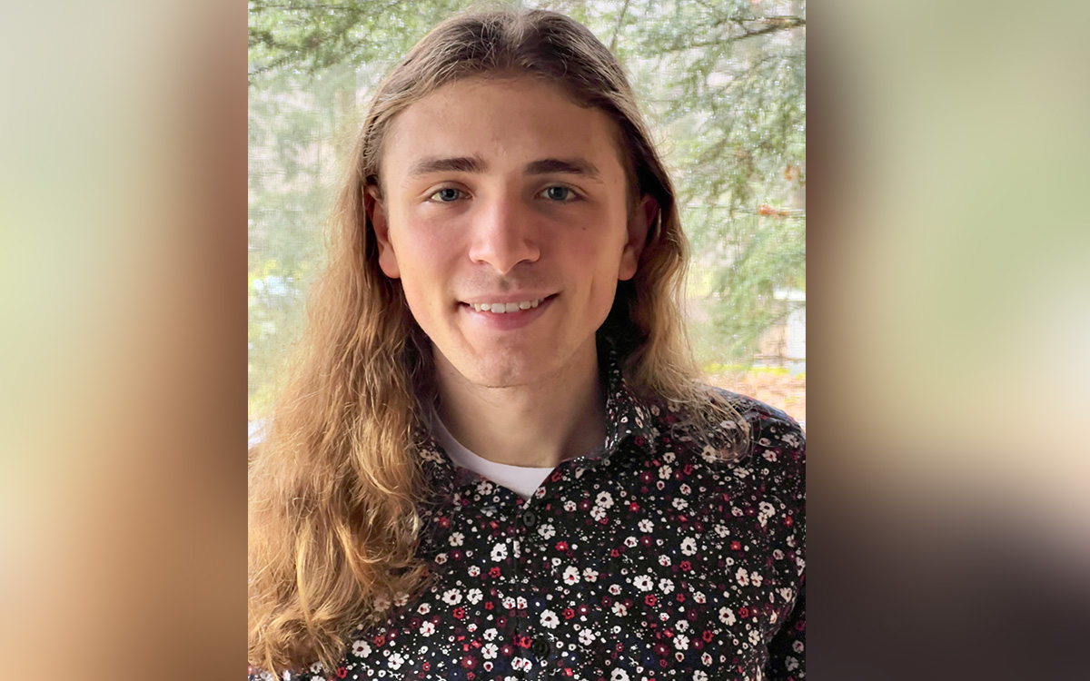 A young man with long blonde hair wearing a floral patterned shirt, smiling in front of a natural background.