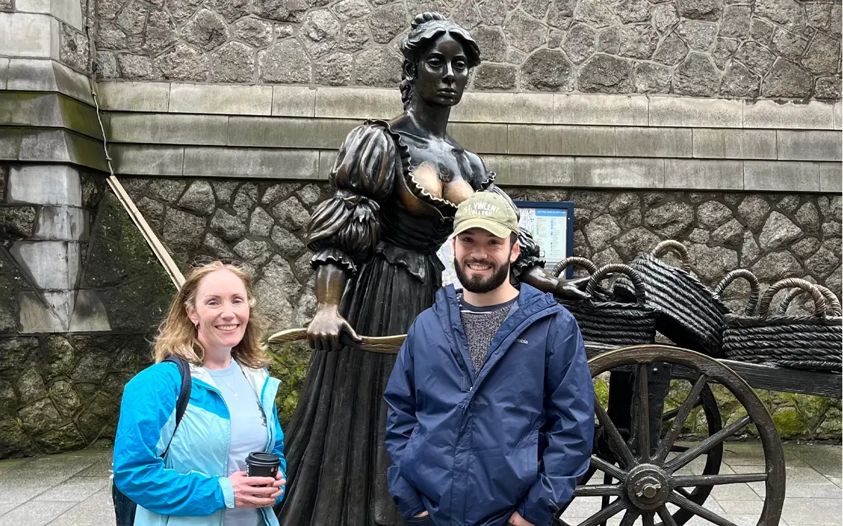 A man and woman pose beside a bronze statue of a woman with a cart, set against a stone wall.