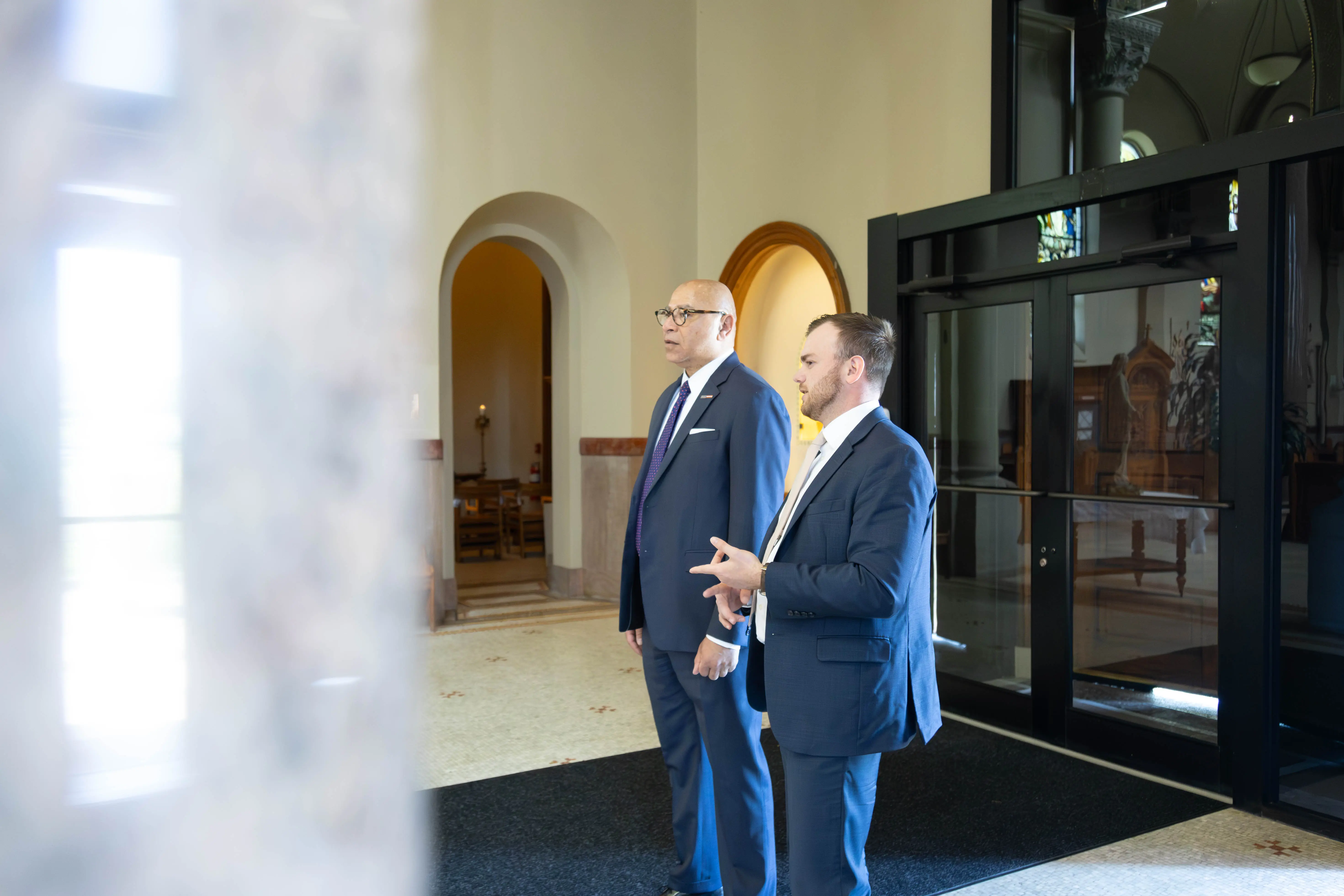 Two professional men in suits standing indoors, engaged in conversation near an entrance with large windows.