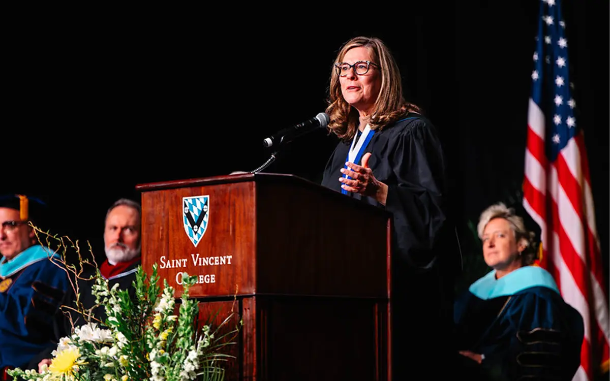 A speaker delivering a speech at the Saint Vincent College graduation ceremony, with faculty members seated on stage and an American flag in the background.