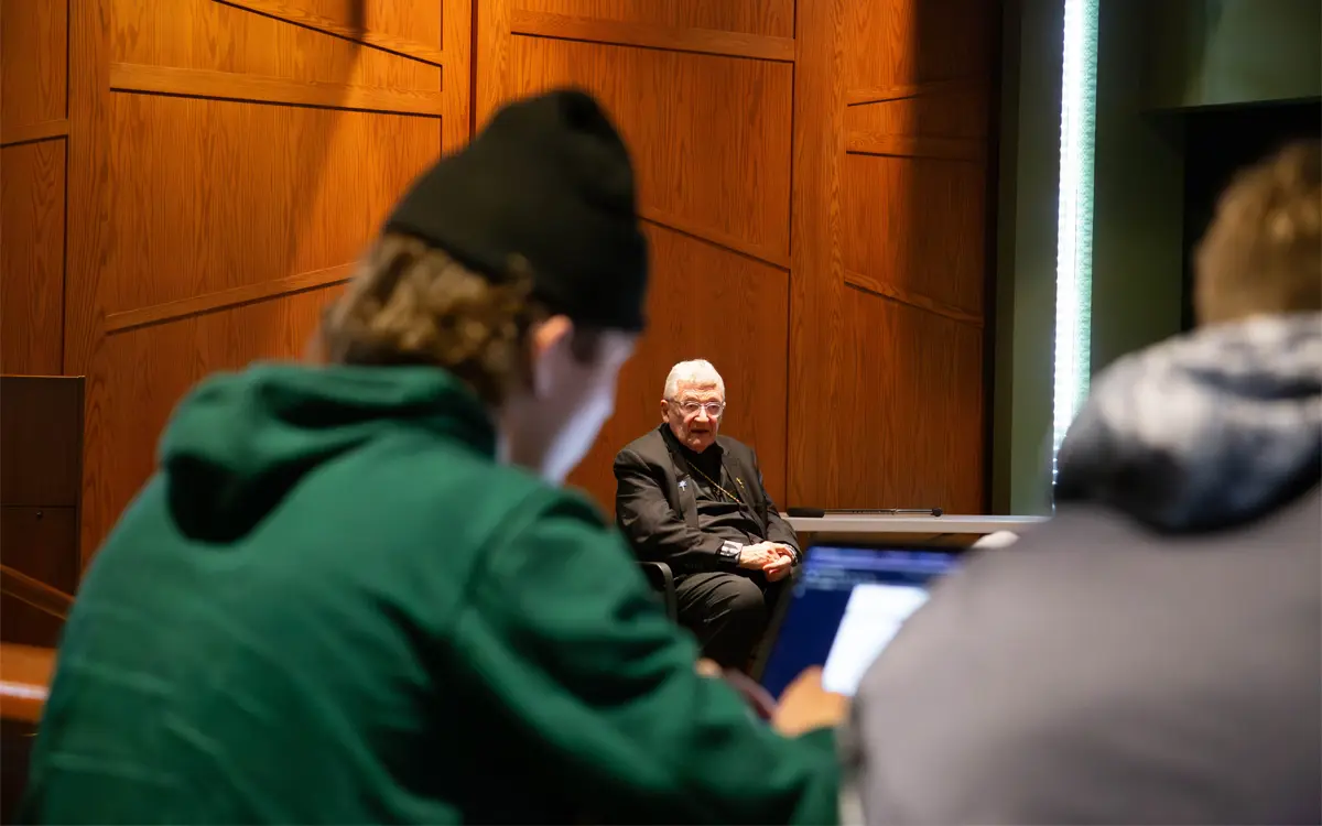 A senior man speaks during a discussion while two students listen attentively, one engaged with a laptop.